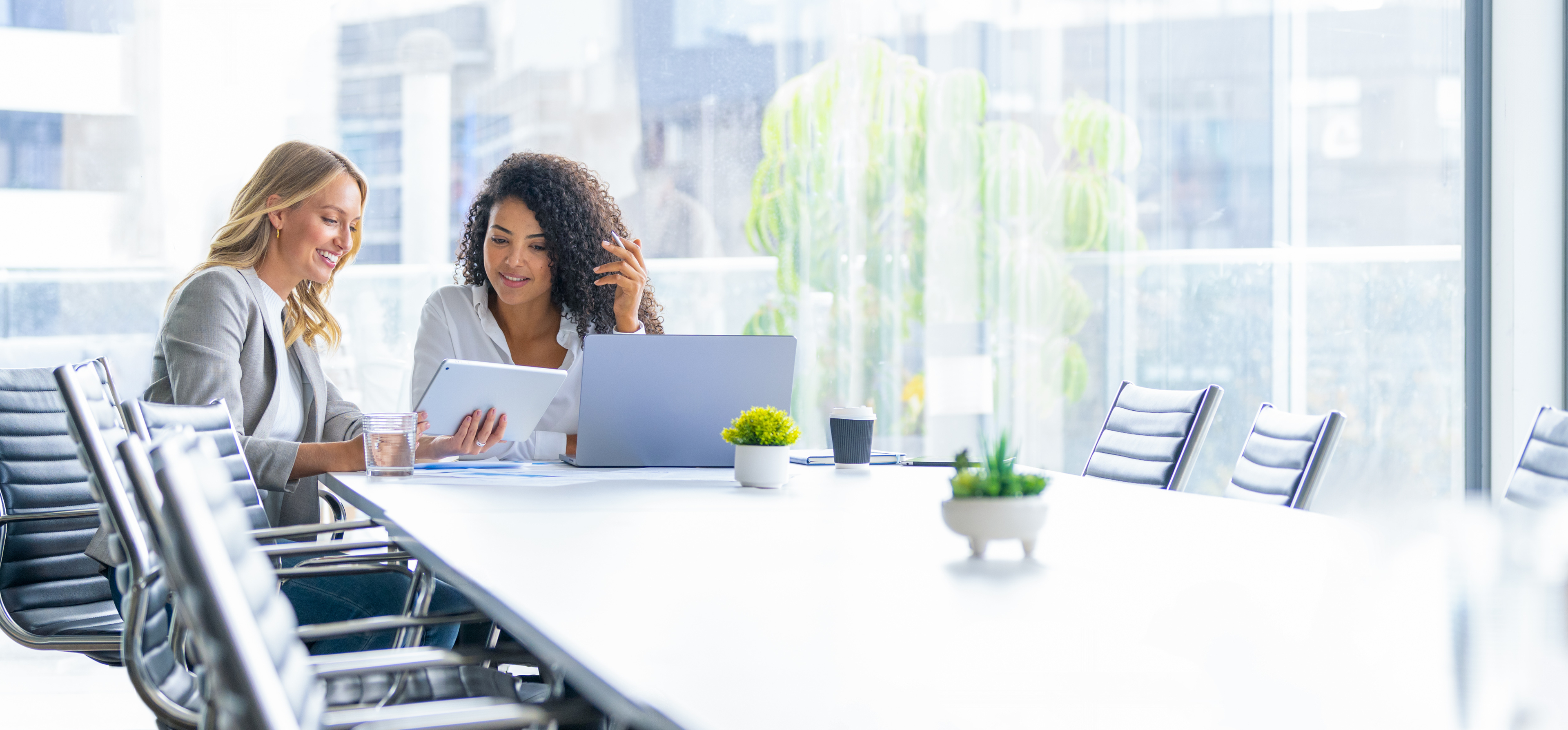 Two professional women sitting at a conference table, reviewing content on a tablet and laptop in a bright modern office
